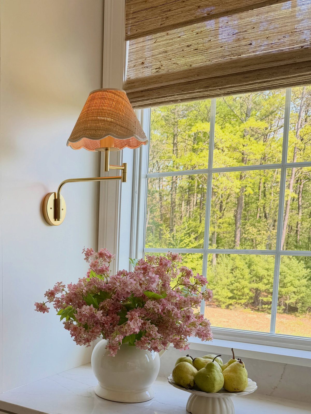 Gold metal wall sconce with a fabric shade mounted next to a window with flowers.