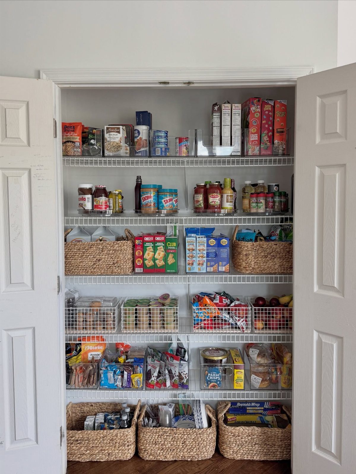 A white pantry closet with wire shelves containing various food items and woven wicker baskets.