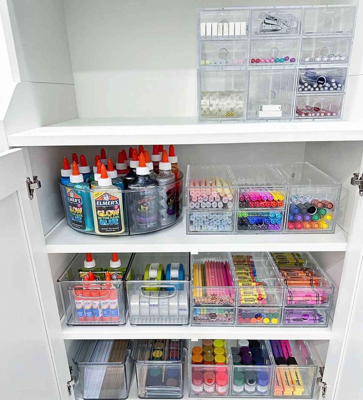 Clear plastic organizers filled with color-coded art supplies on white shelves inside a cabinet.