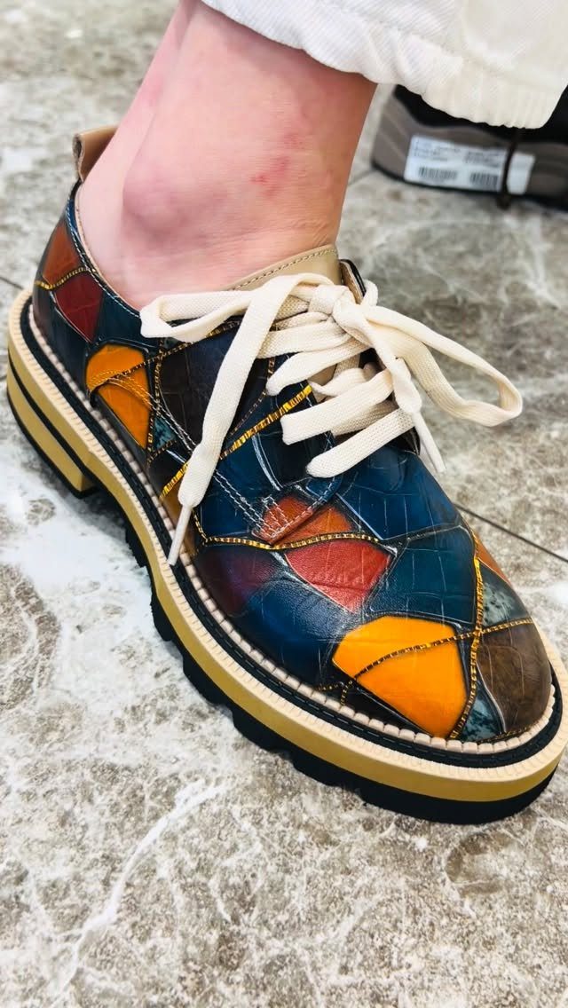 A person wearing colorful patchwork leather derby shoes with cream laces on a textured ground.