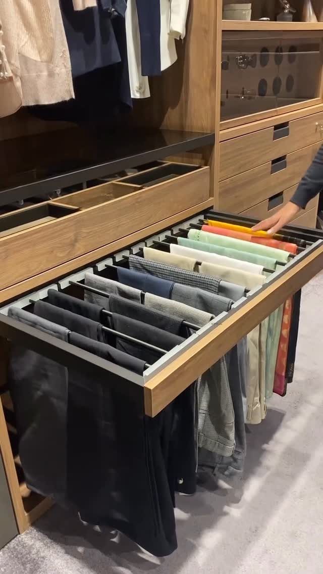 A wooden wardrobe with an open pull-out rack holding several pairs of folded trousers.