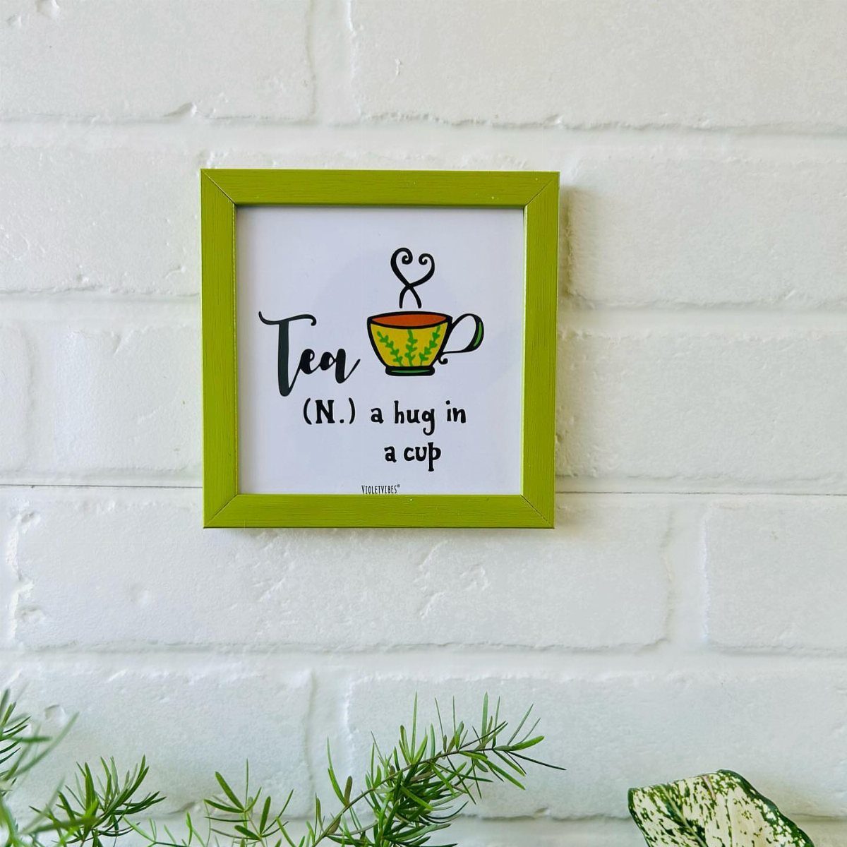 A lime green framed tea quote hanging on a white brick wall above green plants.