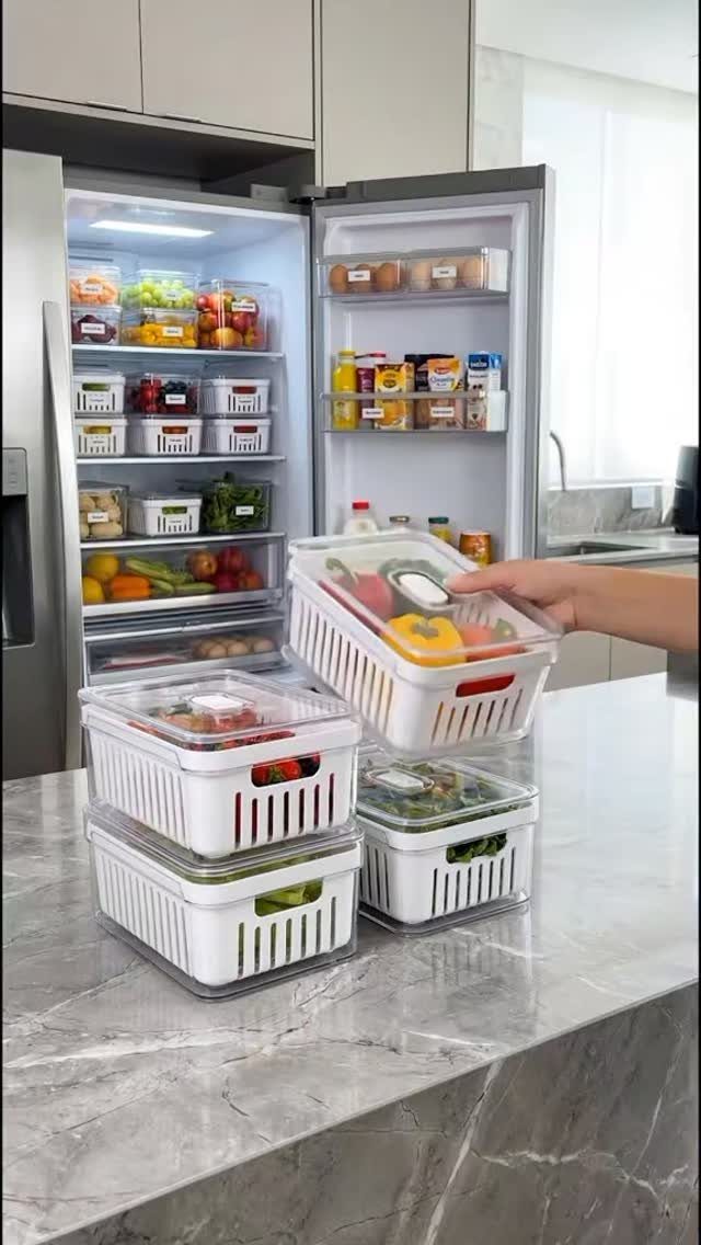 Clear plastic storage bins with white handles being organized on a marble countertop near an open refrigerator.