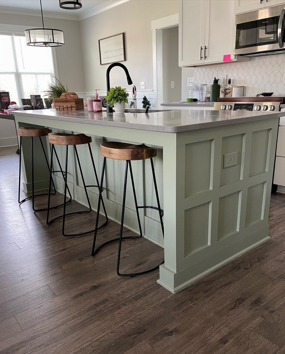 Sage green kitchen island with a grey countertop, black faucet, and wooden bar stools.