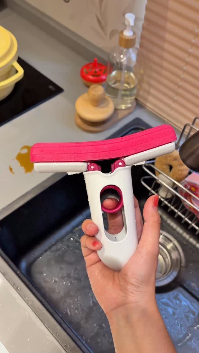 A hand holding a pink and white squeegee tool over a soapy kitchen sink.