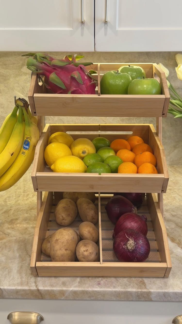 A three-tier wooden organizer filled with various fresh fruits and vegetables on a marble kitchen counter.
